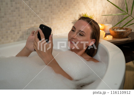 Woman relaxing in a bubble bath at night. She is using her smartphone to browse the internet or watch a video. The bathroom is lit by candles and there is a plant in the background. 116110912