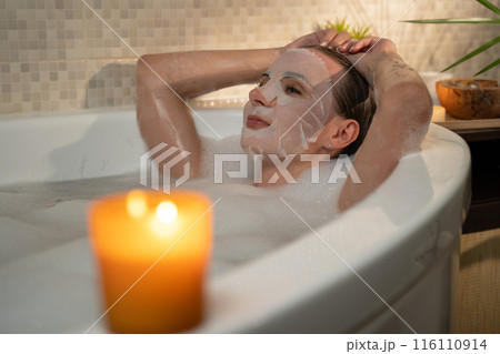 A woman releases in a bubble bath and applied a sweet face mask to her skin. She enjoys a moment of self-care and pampering in her bathroom. 116110914