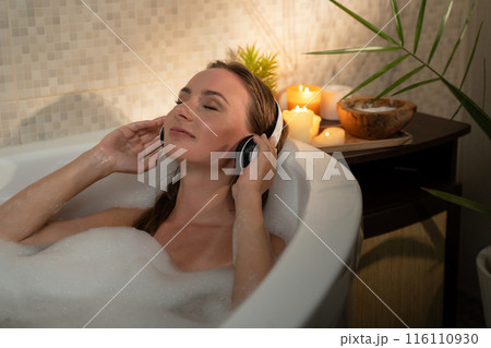 A woman is enjoying a relaxing evening in a bubble bath. She is listening to music with headphones on. The bathroom is dimly lit with candles, and the woman looks serene and content. 116110930
