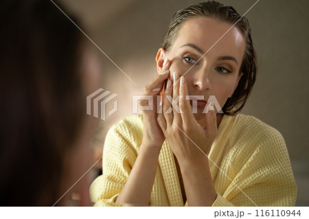 Woman looking in a mirror, focusing on the pimples on her face. She looks concerned and worried about her skin. 116110944
