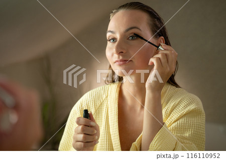 A woman applies mascara to her eyelashes in her bathroom. She is wearing a light yellow robe and looking into the mirror. 116110952