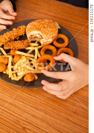 Caucasian man eats half-eaten hamburger, potato chips, onion rings and chicken pieces in gray plate on wooden table 116111178