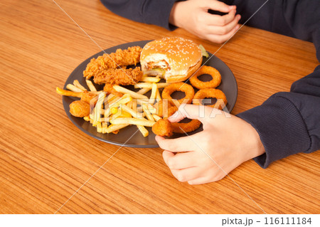Caucasian man eats half-eaten hamburger, potato chips, onion rings and chicken pieces in gray plate on wooden table 116111184