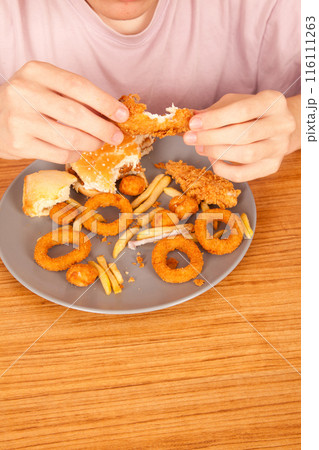 Caucasian man eating fried chicken wings, potato chips, onion rings and chicken wings by hand from a gray plate on a wooden table Caucasian man eating fried chicken wings, potato chips, onion rings and chicken wings by hand from a gray plate on a wooden table 116111263