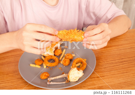 Caucasian man eating fried chicken wings, potato chips, onion rings and chicken wings by hand from a gray plate on a wooden table 116111268