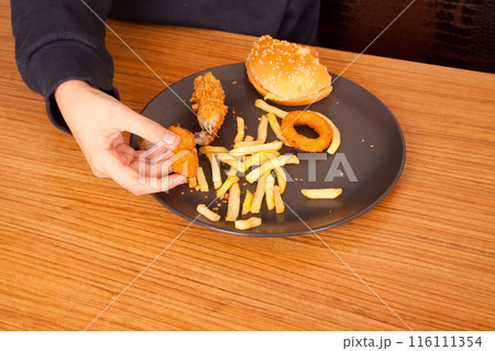 Caucasian man eating fried chicken wings, potato chips, onion rings and chicken wings by hand from a gray plate on a wooden table 116111354