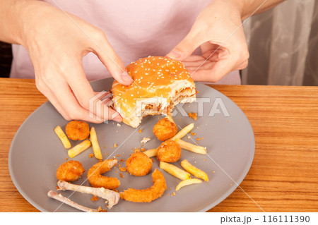 American fast food culture, half-eaten hamburger, potato chips, onion rings and chicken pieces in gray plate on wooden table American fast food culture, half-eaten hamburger, potato chips, onion rings and chicken pieces in gray plate on wooden table 116111390