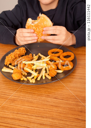 Caucasian man eats half-eaten hamburger, potato chips, onion rings and chicken pieces in gray plate on wooden table 116111442