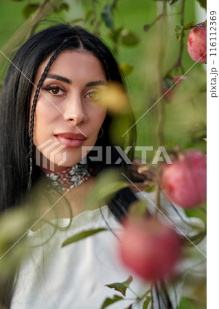 Attractive Caucasian beauty with vivid ethnic necklace posing in apple orchard. Portrait of pretty, young female with little braid, hiding behind apple branch in fall garden. Female beauty concept. 116112369