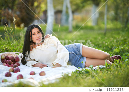 Carefree dark-haired beauty resting on ground, surrounded by scattered apples at sunny day. Side view of happy young woman lying on white rug in early autumn garden. Picnic, beauty concept. Carefree dark-haired beauty resting on ground, surrounded by scattered apples at sunny day. Side view of happy young woman lying on white rug in early autumn garden. Picnic, beauty concept. 116112384