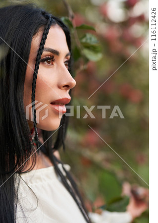 Beautiful dark-haired woman with hairstyle in apple orchard looking to side. Close up profile of attractive Caucasian female with braid, against blurred natural background. Concept of female beauty. 116112426