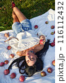 Relaxed pretty woman lying with apples on white rug on grass in garden. View from above of calm Caucasian woman in casual clothes resting on ground, surrounded by apples at sunny day. Picnic concept. 116112432