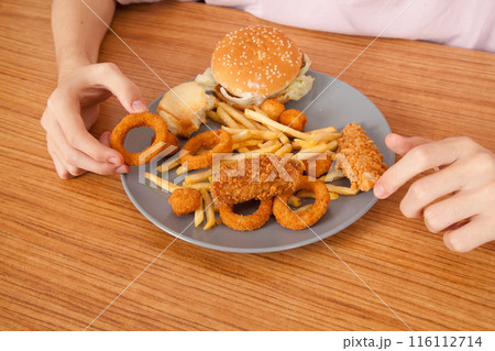 Caucasian man eating fried chicken wings, potato chips, onion rings and chicken wings by hand from a gray plate on a wooden table 116112714