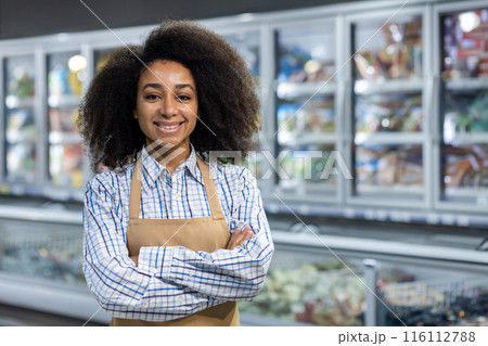 A cheerful supermarket employee wearing an apron and plaid shirt stands confidently with arms crossed in front of the refrigerated section. She exudes friendliness and professionalism A cheerful supermarket employee wearing an apron and plaid shirt stands confidently with arms crossed in front of the refrigerated section. She exudes friendliness and professionalism 116112788