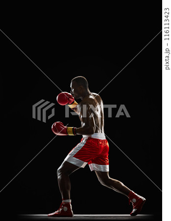 Concentrated African-American man, boxer with sweating shirtless body training, punching isolated on black background. Side view 116113423