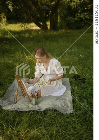 a beautiful young artist in a white dress paints a picture on the river bank a beautiful young artist in a white dress paints a picture on the river bank 116115031