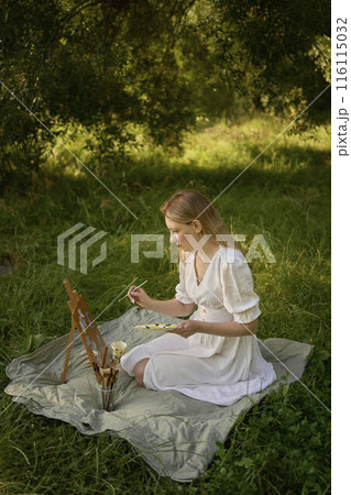 a beautiful young artist in a white dress paints a picture on the river bank 116115032