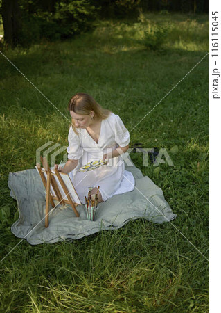 a beautiful young artist in a white dress paints a picture on the river bank 116115045