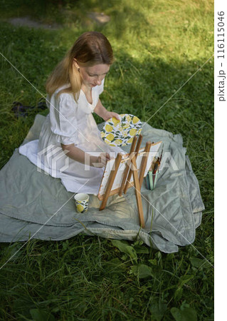 a beautiful young artist in a white dress paints a picture on the river bank 116115046