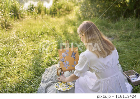 a beautiful young artist in a white dress paints a picture on the river bank 116115424