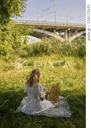 a beautiful young artist in a white dress paints a picture on the river bank 116115431