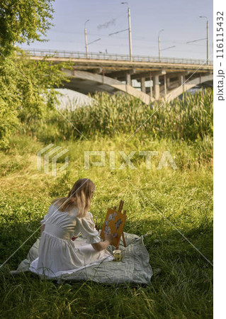 a beautiful young artist in a white dress paints a picture on the river bank 116115432