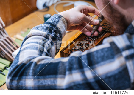 man artisan carpenter carves pattern on wooden casket with a chisel in workshop 116117263