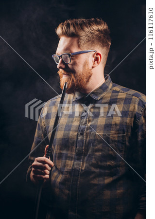 Stylish beard man in glasses and checked shirt smoking hookah at night bar. 116117610