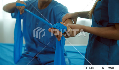 Close up hand patient doing stretching exercise with a flexible exercise band and a physical therapist hand to help in clinic room. Close up hand patient doing stretching exercise with a flexible exercise band and a physical therapist hand to help in clinic room. 116119001