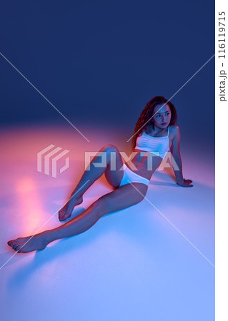 Beautiful young girl with fit, slim body shape, curly long hair, posing in white underwear against blue studio background in neon light Beautiful young girl with fit, slim body shape, curly long hair, posing in white underwear against blue studio background in neon light 116119715