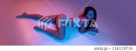 Sensual and attractive young girl with fit body posing on floor in underwear against against blue studio background in neon light Sensual and attractive young girl with fit body posing on floor in underwear against against blue studio background in neon light 116119716