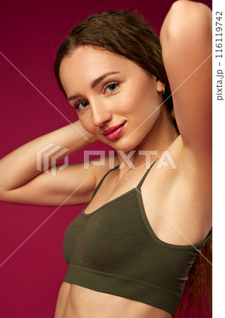 Portrait of attractive young woman with smooth, well-kept skin and nude makeup, posing in green underwear against magenta studio background 116119742