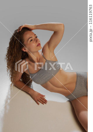 glow of health and well-being. Portrait of young beautiful woman with curly hair and slim fit body, posing in underwear against grey studio background 116119763