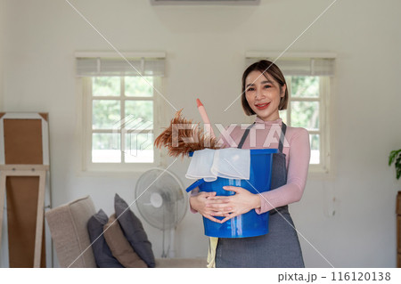 Housekeeper Smiling While Holding Cleaning Supplies in a Modern Living Room Housekeeper Smiling While Holding Cleaning Supplies in a Modern Living Room 116120138