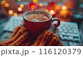 Hands Holding a Red Mug of Hot Drink With Steam in Front of a Computer Keyboard at Night 116120379