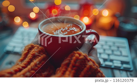 Hands Holding a Red Mug of Hot Drink With Steam in Front of a Computer Keyboard at Night 116120379