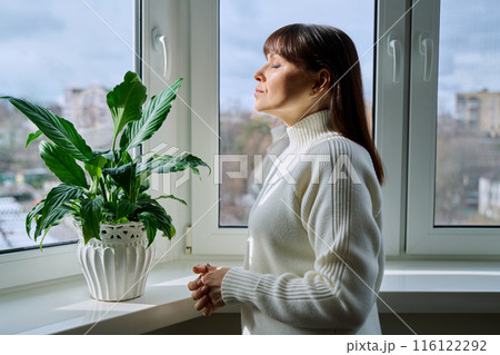 Middle aged woman with closed eyes enjoying winter sun at home near window Middle aged woman with closed eyes enjoying winter sun at home near window 116122292
