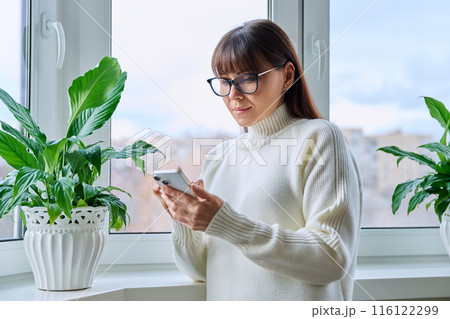 Calm confident middle aged woman using smartphone, at home near window 116122299