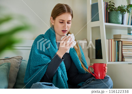 Young sick woman sitting under warm blanket sneezing coughing in handkerchief 116122338