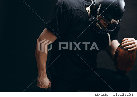 American football player with helmet and armour with a ball against black wall 116123152