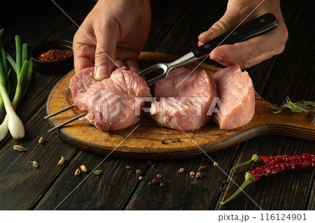 The cook hands preparing a meat steak for grilling. Chef hands skewering raw veal meat with a fork on the kitchen counter. Advertisement space on black table 116124901