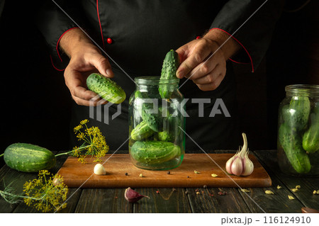 Chef hands filling jar with cucumbers before canning with spices on kitchen table. Concept of pickling tasty cucumber Chef hands filling jar with cucumbers before canning with spices on kitchen table. Concept of pickling tasty cucumber 116124910