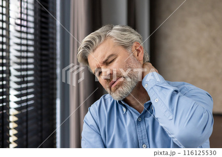 A middle-aged businessman in a blue shirt experiences neck pain while sitting in an office. The image depicts discomfort and stress, common feelings for office workers. A middle-aged businessman in a blue shirt experiences neck pain while sitting in an office. The image depicts discomfort and stress, common feelings for office workers. 116125530