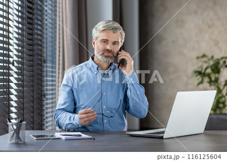 Mature businessman with gray hair holding glasses, talking on phone at office desk with laptop and notepad. Modern office setting with blinds and plant. 116125604