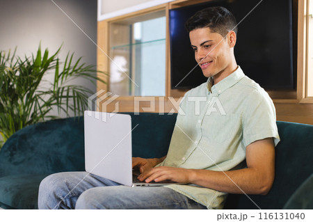 Biracial young man using laptop on blue sofa in a modern business office 116131040
