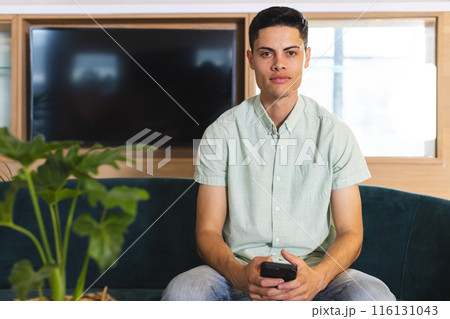 A young biracial man holding a smartphone sits on a couch in a modern business office A young biracial man holding a smartphone sits on a couch in a modern business office 116131043