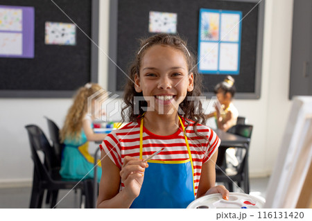 In school art class, biracial young girl holding a paintbrush smiles at camera 116131420