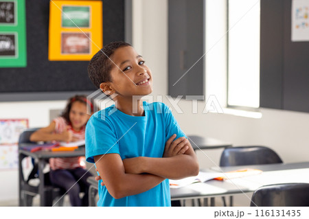 In school, young biracial boy standing with arms crossed, looking thoughtful in the classroom 116131435