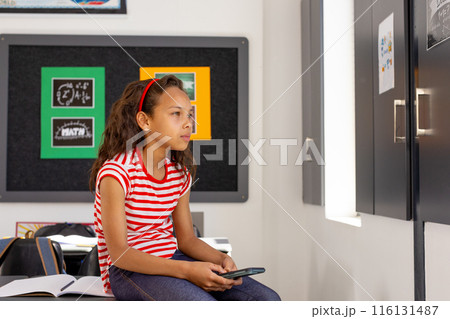 In school, in classroom, young biracial girl holding a smartphone looks thoughtful In school, in classroom, young biracial girl holding a smartphone looks thoughtful 116131487