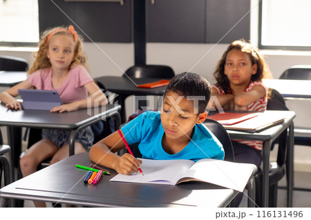 In school, in classroom, boy in blue shirt writing in notebook, two girls watching 116131496